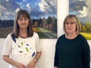 Evi Steiner-Böhm und Gabriele Kastner von der Regionalbibliothek Weiden stehen vor zwei Landschaftsbildern in Öl gemalt und lächeln in die Kamera